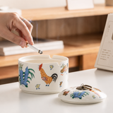 Hand holding cigarette above lidded ceramic ashtray with rooster and chick pattern