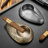 Vintage engraved alloy cigar ashtrays in antique gold and gunmetal silver with cigars on tabletop