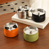 Four short stainless steel ashtrays with rotatable lid, in orange, green, silver, and black on a wooden surface.