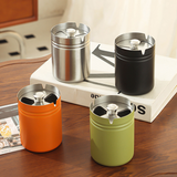 Four cylindrical stainless steel ashtrays with rotatable lid, in orange, green, silver, and black on a wooden surface.