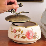Decorative ceramic ashtray with floral designs and a bronze lid on a wooden surface.
