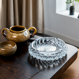 Clear glass ashtray on a wooden table with a ceramic teapot and bowl in the background.