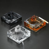 Clear, gray, and amber square glass ashtrays with cigarette rests on dark surface