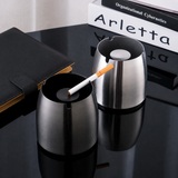 Two silver stainless steel ashtrays with center snuffer plates on a black table