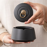 Hand displaying the removable lid above a black ceramic ashtray