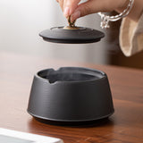 Hand lifting the black ceramic ashtray lid using the metal ring handle