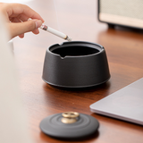 Hand holding a cigarette over an open black ceramic ashtray with lid nearby