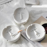 White face expression ceramic ashtrays styled on soft fabric, elegant tabletop scene
