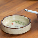Round cream ceramic ashtray holding a lit cigarette, with speckled glaze and floral-style design