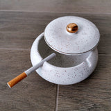 White speckled covered ceramic ashtray with gold knob and cigarette on a wooden tabletop