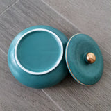 Open teal ceramic ashtray with matching gold-knob lid on a wooden tabletop